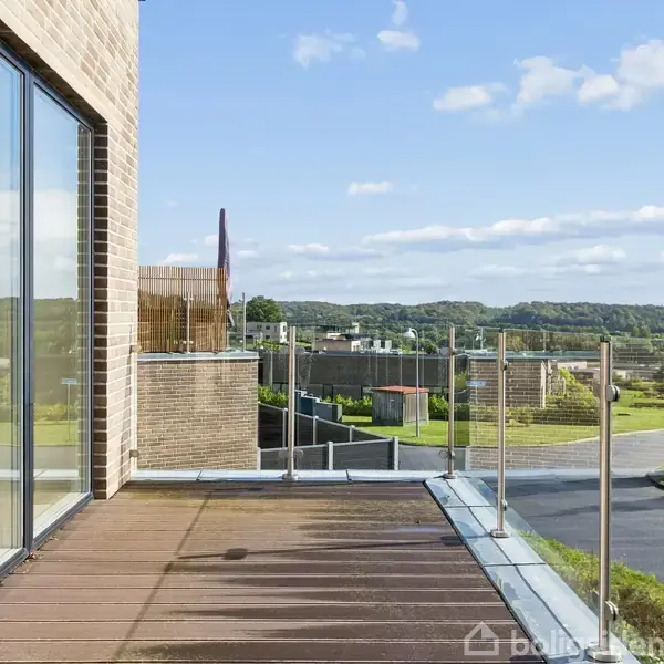 En terrasse med glasrækværk og trædæk vender ud mod en naturskøn udsigt med bakker og spredte huse i baggrunden. En stor glasfacade reflekterer omgivelserne.