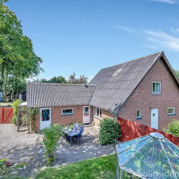 Et rødt murstenshus med grått tag står omgivet af en have med planter og en glasoverdækket terrasse. Der står havemøbler på belagt grund foran huset, bag et rødt træhegn.