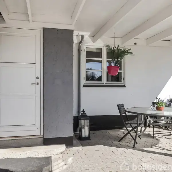 Dør til entré ved overdækket terrasse. Bord med stole foran vindue. Grydeplanter hængende fra loftet. Udemøbler og gril i baggrunden. Flisebelagt terrasse.