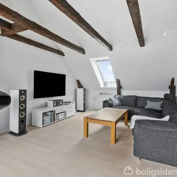 Guitar hænger på væggen i en stue med skrå loftsbjælker. Væghængt pejs brænder ved siden af et TV og højttalere. Grå sofa og trægulv giver rummelig indretning.