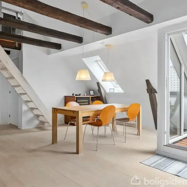 Et lyst rum med et spisebord og tre stole i forgrunden. En lænestol står ved siden af glasdøre, der åbner til en balkon med grill. En guitar hænger på væggen.