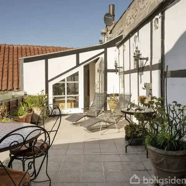 Terrasse med to liggestole vendt mod hvide bindingsværksvægge med sorte bjælker, omgivet af planter i krukker og et rundt bord med metalstole på terrassens flisebelagte gulv.