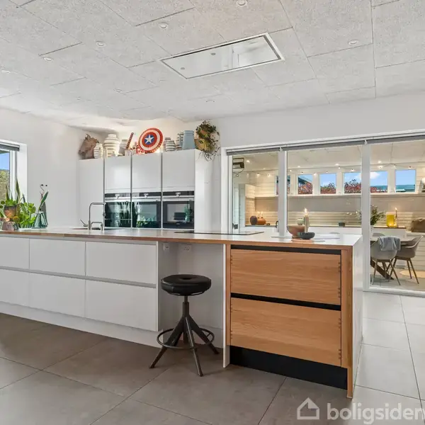 Et moderne køkken med en stor ø, barstol og hvide skabe. Der er glasdøre til en spisestue, og planter pynter rummet. Et Captain America-skjold er udstillet ovenpå skabene.