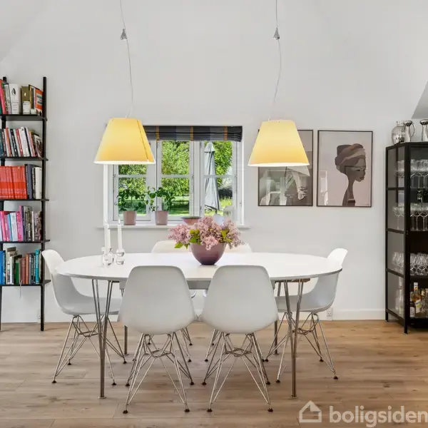 Et spisebord med seks hvide stole står i en lys spisestue. To lamper hænger over bordet, omkranset af en bogreol og et vitrineskab. Blomster pryder bordet.