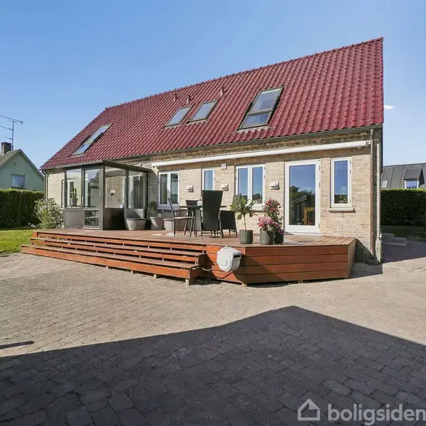 Stor, grå sofa står ved en stue med træbord. Bordlampe og pyntepuder tilføjer hygge. Lysindfald fra flere vinduer. Væggene har kunstværker, og en lænestol står i hjørnet.