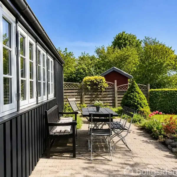 Sort træhus med hvide vinduer, langbord og stole på flisebelagt terrasse. Haven er omkranset af grønne buske og et lille, rødt skur i baggrunden under en klar blå himmel.