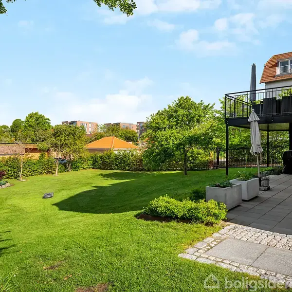 En velplejet græsplæne breder sig i en solrig have med træer og buske. En terrasse med stole og en parasol ligger ved et hus med rødt tag i baggrunden.