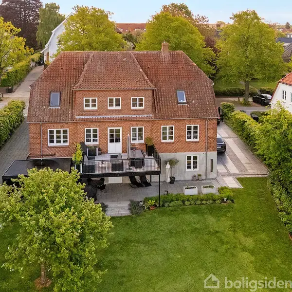 Rødt murstenshus med rødt tegltag, omgivet af grøn have med hække. To etager med kvistvinduer, terrasse foran huset, placeret i et villakvarter med andre lignende boliger og træer.
