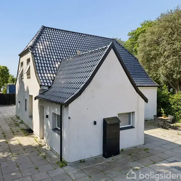 Et hvidt, moderne hus med sort tegltag står stille på flisebelagt terrasse. Der er træer og buske rundt om, som skaber en grøn og fredelig atmosfære.