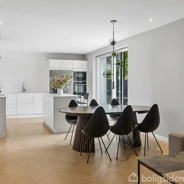 Et moderne køkken-alrum med et sort spisebord og seks sorte stole. Køkkenet har hvide skabe, og der er to store døre, der fører ud til en terrasse.