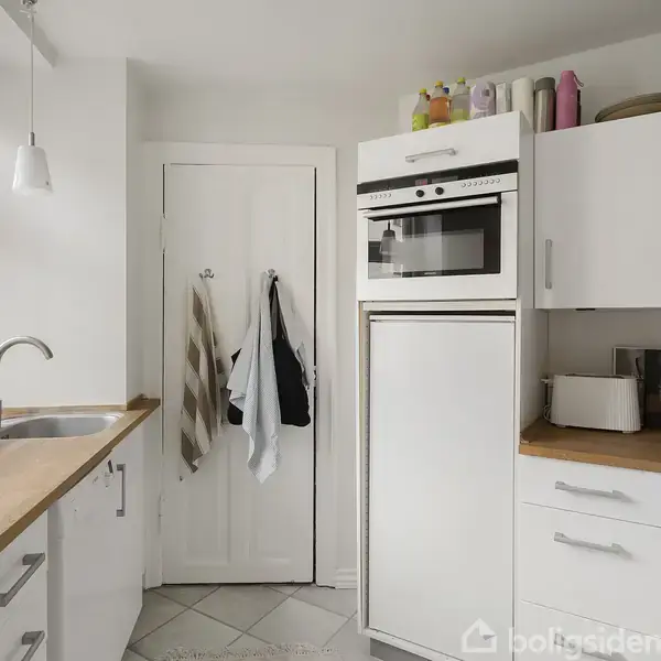 Et hvidt køkken med træbordplader, hvor en indbygget ovn og høj køleskab står langs væggen. En rød blender og køkkenredskaber ses på bordet. Håndklæder hænger på døren.