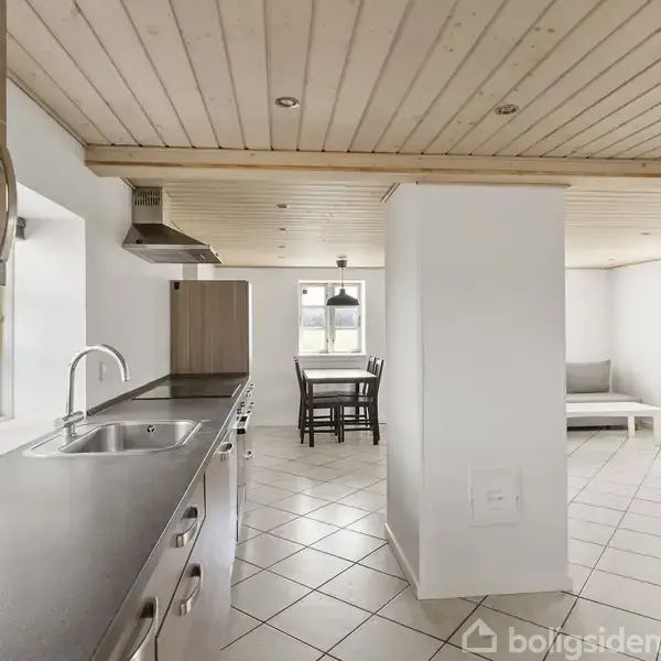 Køkken med sølvvask og køkkenbord, åbner op mod spiseområde med bord og stole. Lyse fliser på gulvet, træloft og lys fra vinduer.