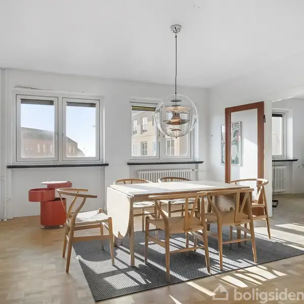 Spisebord med seks stole står i et lyst rum. En moderne lampe hænger over bordet. Store vinduer og en skydedør med glas åbner til et tilstødende rum med en plante.