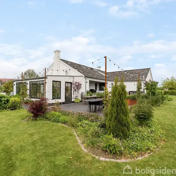 Et hvidt hus med gavltag står omgivet af en velplejet have. Et terrasseområde foran huset har havemøbler. Grønne buske og træer omkranser området under en blå himmel.