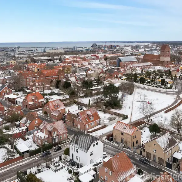 Bybygninger dækket af let sne står i en tæt bymidte med røde tage, omkringliggende veje og skovklædte områder. I baggrunden ses en havn og kystlinje.