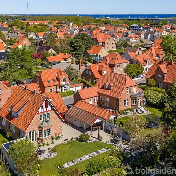 En række røde murstenshuse med røde tegltage ligger i et villakvarter. Husene står tæt i grønne omgivelser med træer og haver, og der er udsigt mod vandet i baggrunden.