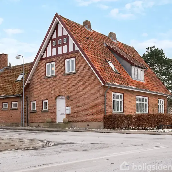Rødt murstenshus med rødt tegltag, trekantet gavl og skorsten. Snebelagt fortov og vej i forgrund. Lygtepæl og busk i nærheden. Blå himmel i baggrunden.