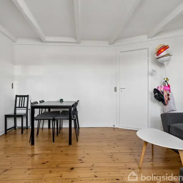 Stue med sort spisebord og fire stole. En grå sofa står ved hvidt sofabord. Trægulv, hvidmalede vægge. Hjelme hænger på kroge nær døren.