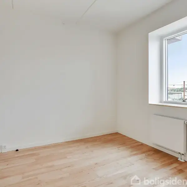Et tomt værelse med trægulv og hvide vægge. Et enkelt vindue lader naturligt lys trænge ind. En radiator er placeret under vinduet i et minimalistisk interiør.