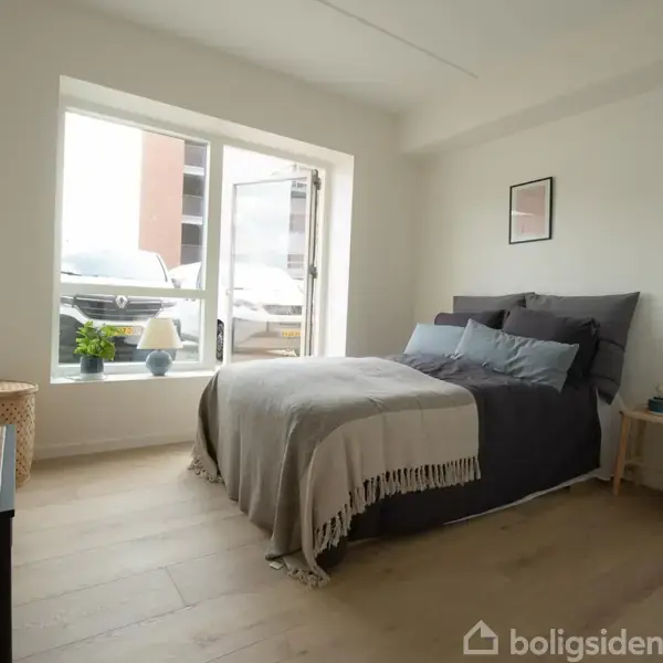 Seng med sengetæppe og puder står i et lyst soveværelse med trægulv. Ved vinduet står en plante og en lampe.