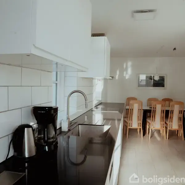 Køkkenbord med kaffemaskine og elkedel, står på en blank bordplade. I baggrunden er der et spisebord med træstole. Lyse fliser og vægge omkranser rummet.