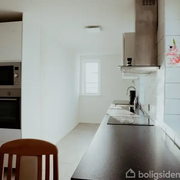 Et moderne køkken med sort bordplade og vask, omgivet af hvide skabe og en indbygget ovn. Et vindue for enden giver naturligt lys.
