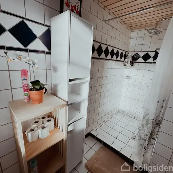 Et badeværelse med bruser er omgivet af hvide fliser og sort diamantmønsterbort. En hvid reol står ved siden af en træreol med orkidé, sprayflaske og toiletruller.