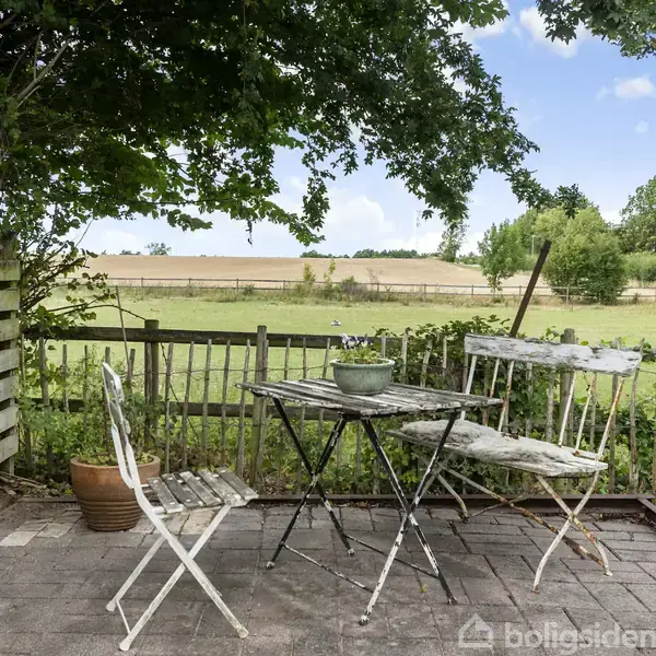To hvide metalstole og et bord med en plante står på en terrasse med flisebelægning. De omkranses af et lavt hegn og naturgrøn udsigt i landlige omgivelser.