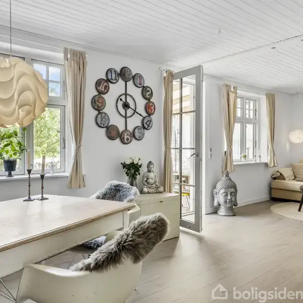 Stue med spisebord og stole dækket med pels, stor urdekoration på væggen, og sofaområde i baggrunden. Store vinduer med lys gardiner giver udsigt til grøn have udenfor.