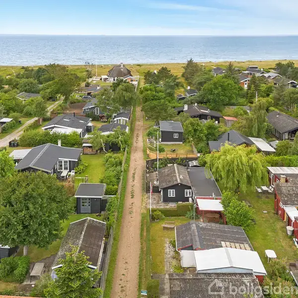 Række af sommerhuse langs en grusvej ved kysten. Husene er omgivet af træer og grønne områder, med havet i baggrunden under en skyfri himmel.