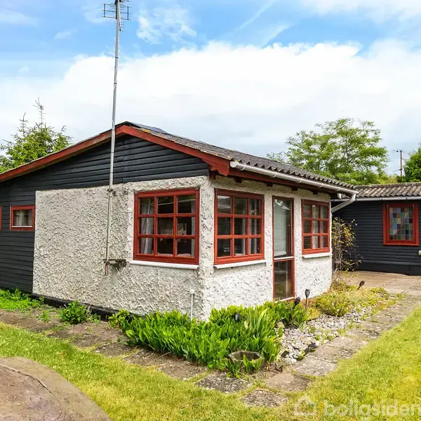 Et lille hus med røde vinduesrammer står stille i en have med grøn græsplæne og blomsterbede. Der er et mindre skur i baggrunden, omgivet af hegn og buske.