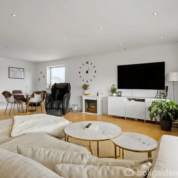 Beige sofa står i en lys stue, ved siden af runde borde. Fjernsyn og planter er på en skænk. Vægur hænger ved spisebord med stole i baggrunden.