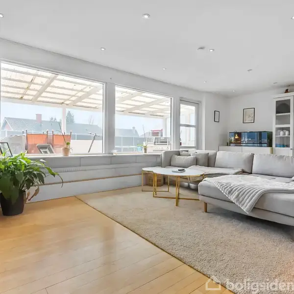 Stor grå sofa står i en moderne stue, beliggende på en lys trægulv. Sofaen er placeret foran store vinduer, som vender ud mod en terrasse, og omgivet af planter og en bogreol.