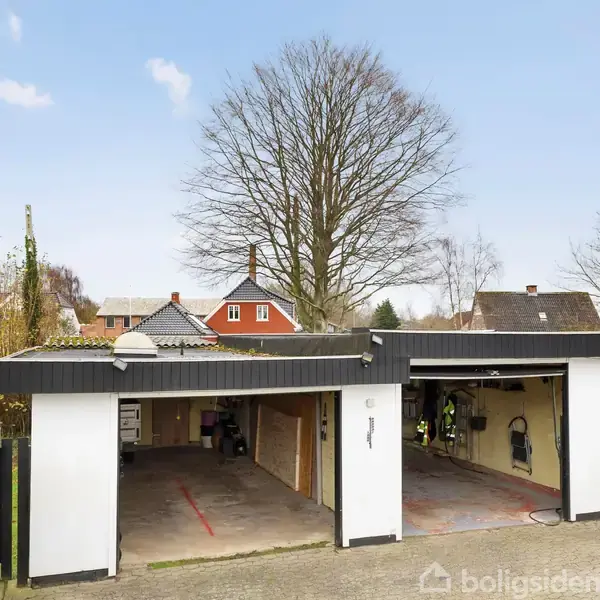 Garage med to åbne porte, stående på en flisebelagt indkørsel. Græsplæne og træer i baggrunden under en klar himmel. Der er ingen tekst i billedet.