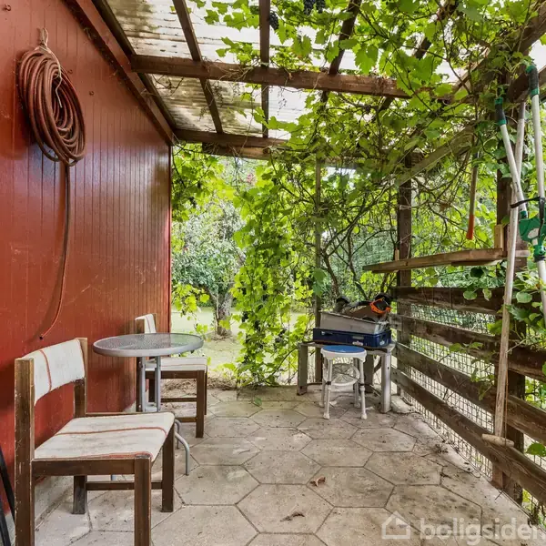 To stole og et lille bord står på en terrasse med pergola overgroet af grønne planter. En rød væg til venstre og en grill nederst til højre i billedet.