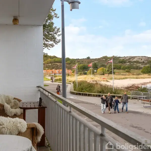 Balkon med stole dækket af fåreskind står mod havudsigt. Mennesker går langs en strandpromenade nær kysten, flankeret af danske flag og grønne bakker i baggrunden.