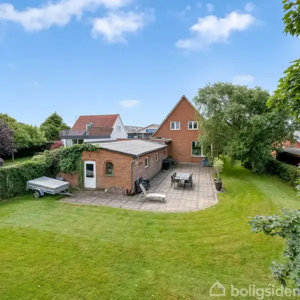 Rødstenshus med terrasse står i en velplejet have. En havemøbelgruppe er placeret på terrassen. Omkring huset er der grønne buske og træer under en klar blå himmel.