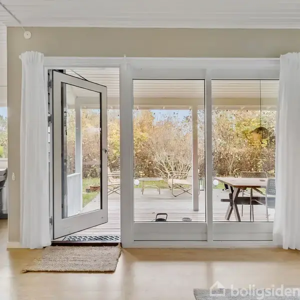 Dør i et moderne hjem står åben mod en terrasse med møbler. Indretningen er lys og luftig med spisebord og stole i baggrunden.