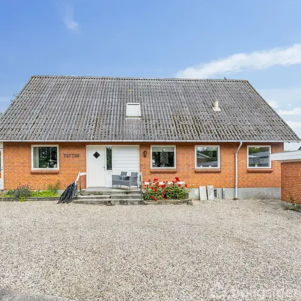 Et rødt murstenshus med asymmetrisk tag står på en gårdsplads med grus. Trappetrin fører til hoveddøren omgivet af blomsterbede. Tekst: "768370". Der er en carport til højre.