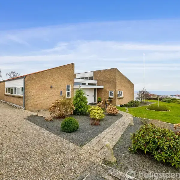 Et moderne hus af mursten med fladt tag står stille omgivet af en flisebelagt indkørsel, grønne buske, og en velplejet græsplæne under en klar blå himmel.