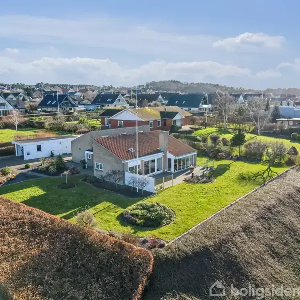 Et hus med rødt tag og udestue står omgivet af velplejede haver og grønne plæner i et boligområde med flere huse i baggrunden under en blå himmel.