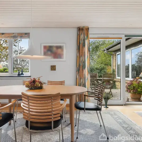 Rundt spisebord med træstole står i et lyst rum med store vinduer og terrassedør. En lampe hænger over bordet, og en terrasse med planter kan ses udenfor.