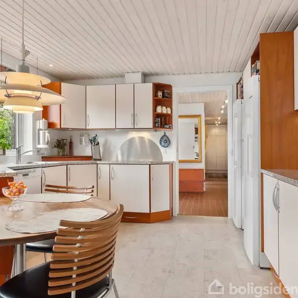 Et moderne køkken med hvidt inventar og trædetaljer. Et rundt spisebord med stole foran et vindue, og en vedhæftet lampe. Køkkenet er lyst med udsigt til en grøn baggrund.