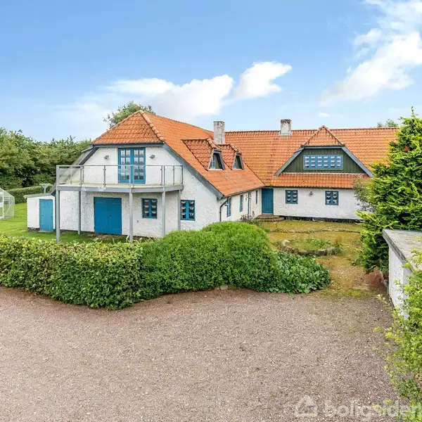 Huset står i fokus med hvid facade og rødt tegltag, omgivet af grønt hegn og have. Der er en indkørsel foran. Vejen fører til skov og natur i baggrunden.