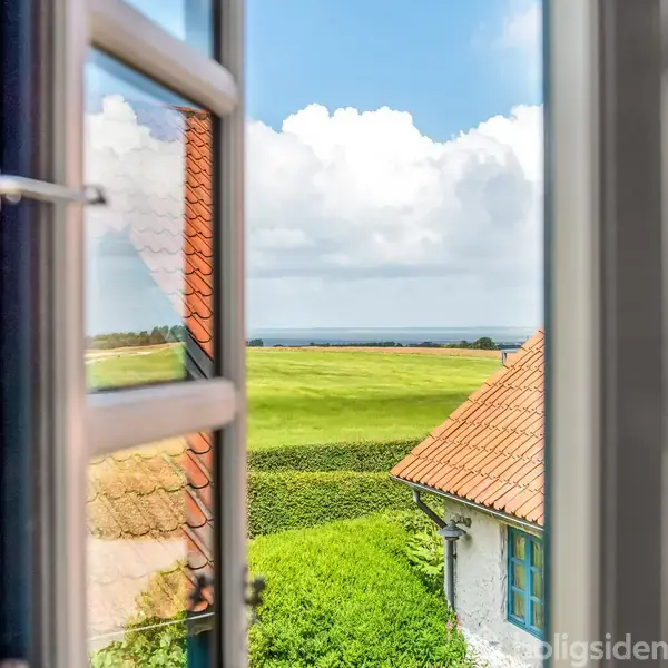 Et åbent vindue viser en udsigt over grønne marker og blå himmel med skyer, omgivet af bygninger med røde tegltage.