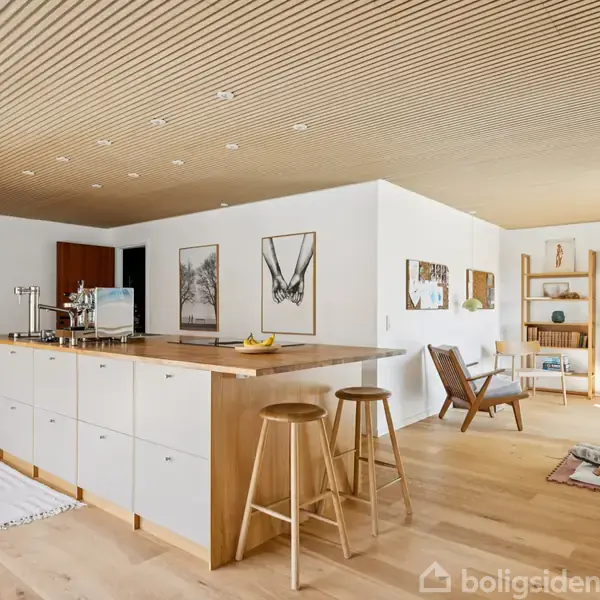 Køkken-ø står i midten med træoverflade og tre barstole. Lysindfald fra store vinduer oplyser stuen med reoler og stole i baggrunden. Træloft og gulve skaber en varm atmosfære.