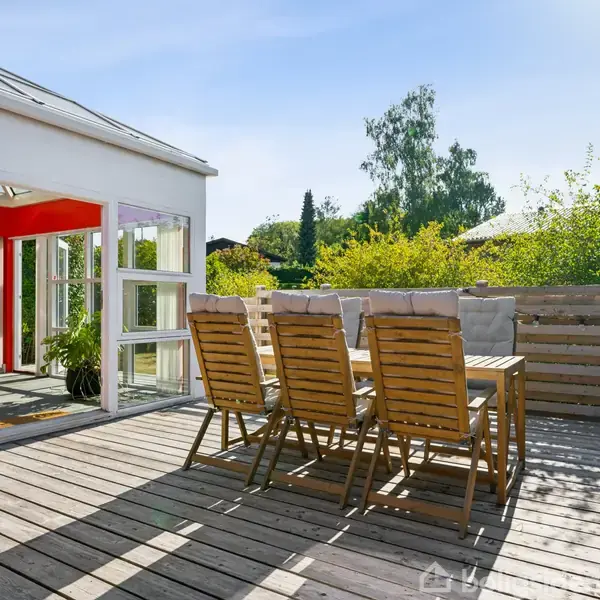 Træstole omkranser et udendørs bord på en solrig træterrasse. Ved siden af er en moderne bygning med store vinduer, omkranset af grønne buske og et træ i baggrunden.