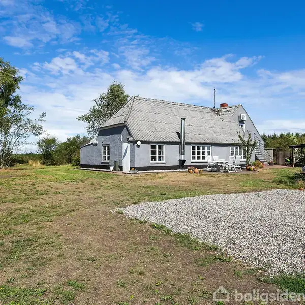 Et hvidt hus med stråtag står stille på en rummelig græsplæne. Omkring huset er der træer og en blå himmel, samt et hvidt grusområde foran.