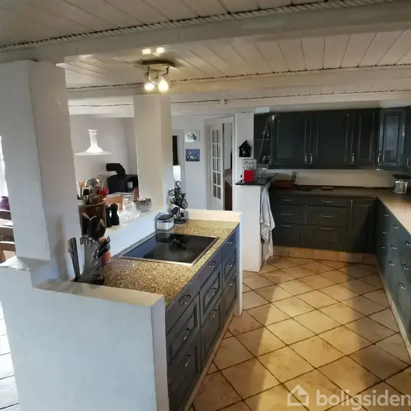 Et køkken med mørke træskabe, der strækker sig langs væggene. En kogeø med køkkenredskaber står i midten. Et spiseområde ses i baggrunden ved et vindue med naturligt lysindfald.