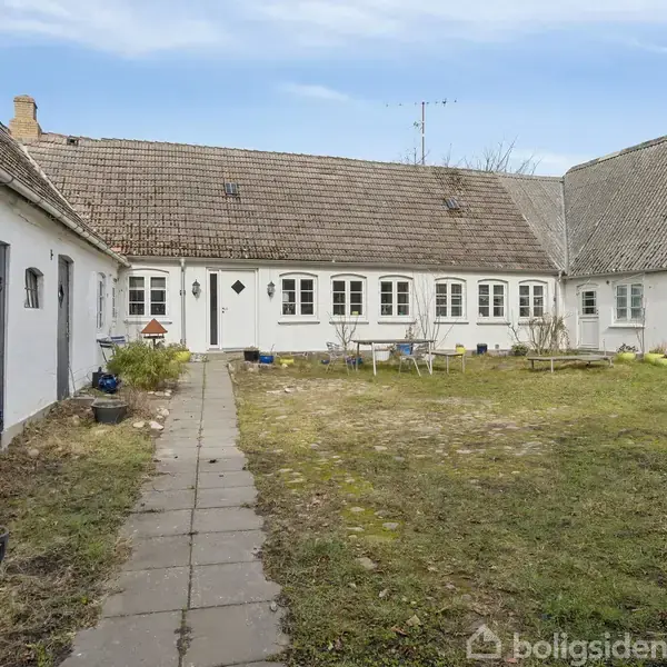 Bindingsværkhus står stille midt i vintertræer, omgivet af en grøn græsplæne. Et tag med mos understreger husets alder. Skoven i baggrunden antyder en landlig atmosfære.